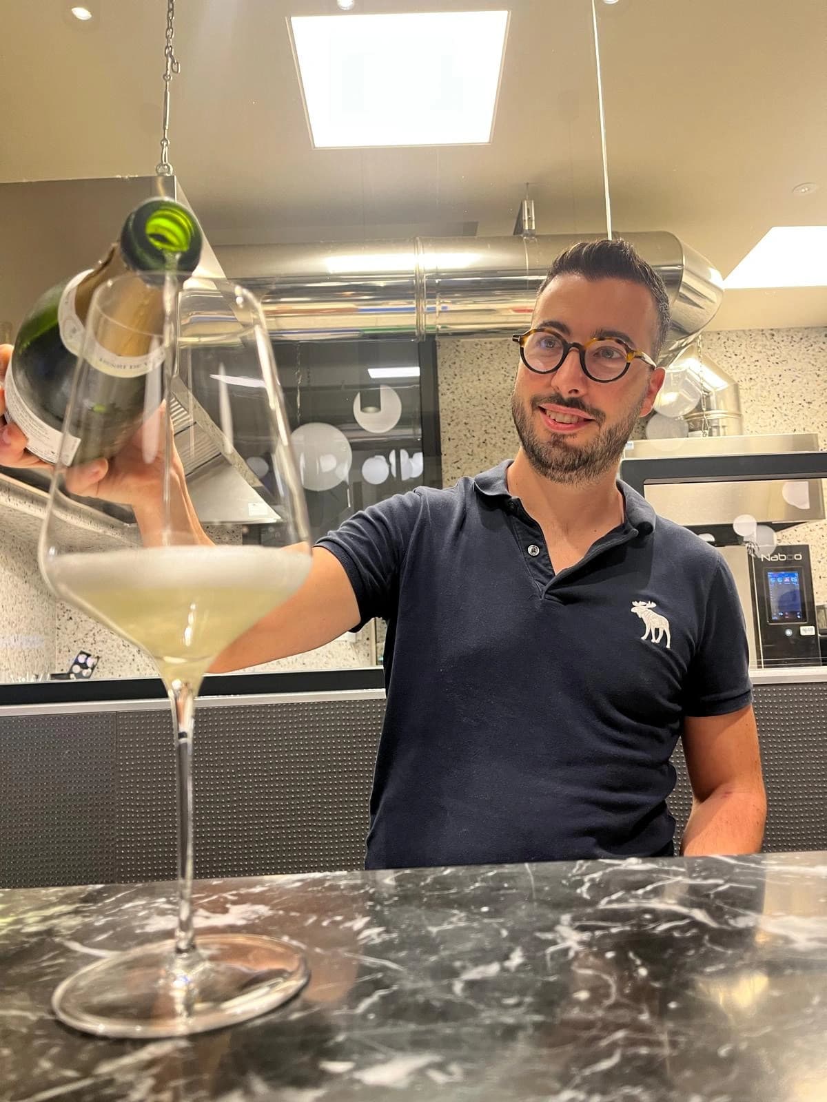 Mattia Raiti versa un vino spumante al bancone di Materia, Osteria Contemporanea, con la cucina a vista alle spalle